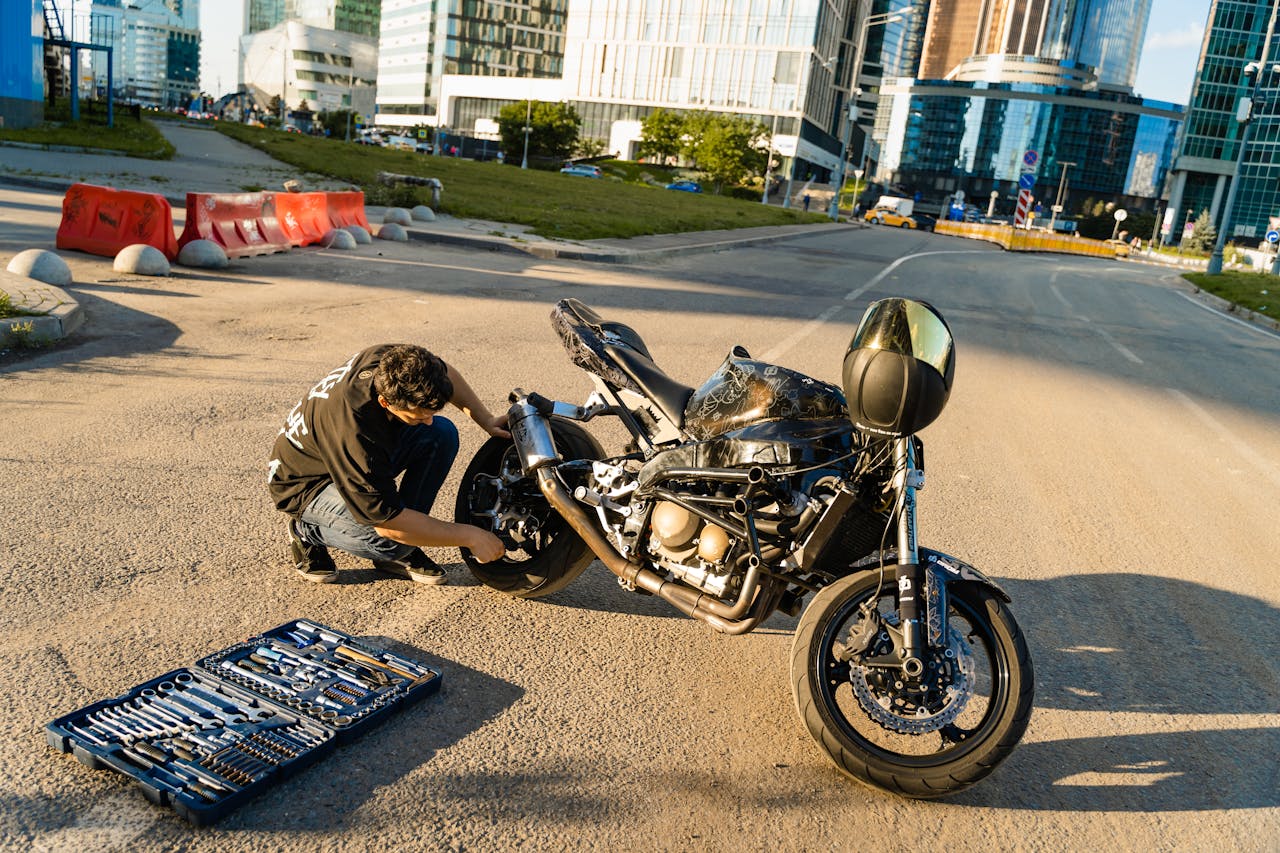 A man repairs his motorcycle on a city street, surrounded by urban architecture, under a bright afternoon sun.