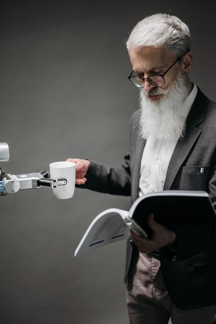 contact-img An elderly scientist reads while a robot serves coffee, representing technology's role.